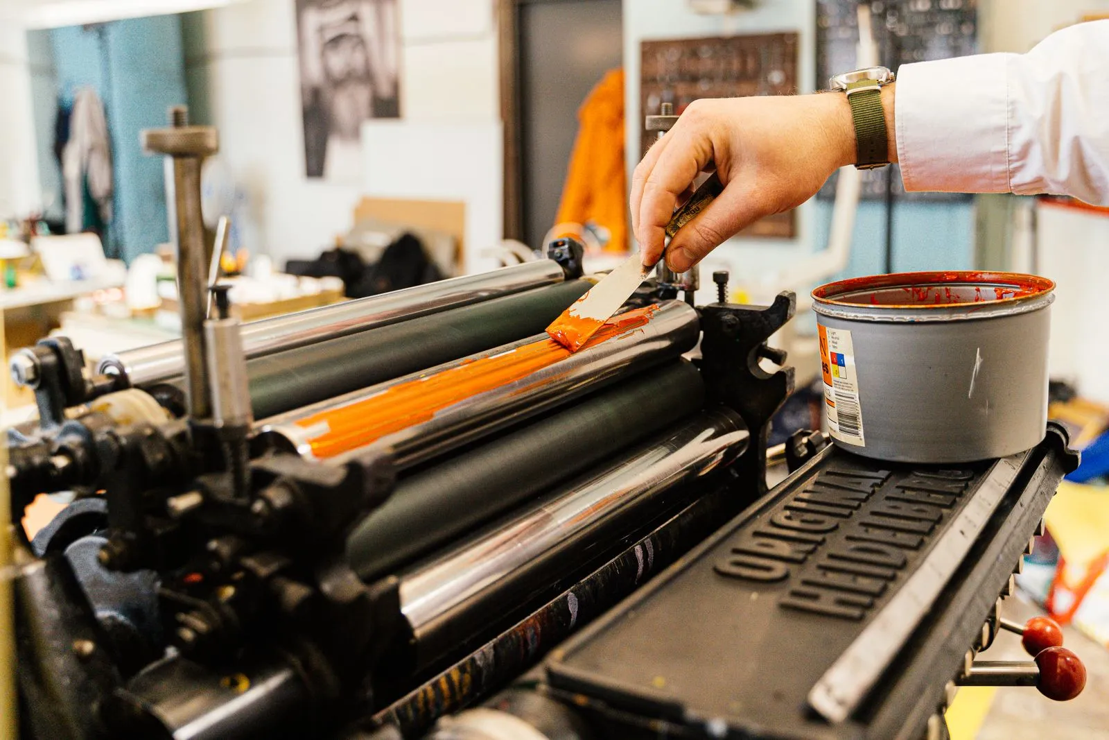 A detailed view of a hand applying orange ink to a vintage printing press roller indoors.